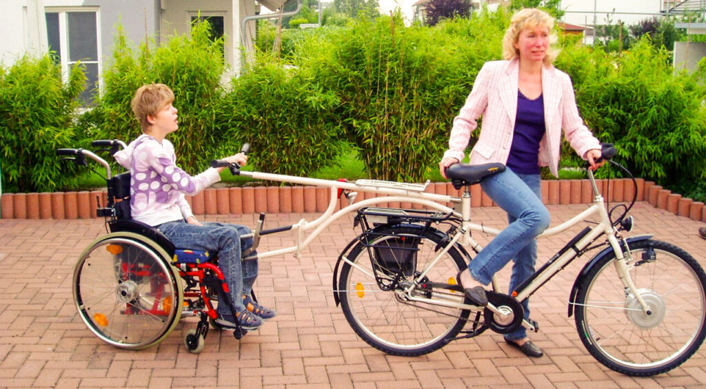 Melanie sitzt in ihrem neuen Rollstuhltandem. Ihre Mutter hält das Zugfahrrad.