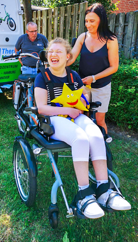 Milena sitzt freudestrahlend in ihrem neuen Elektro-Rollstuhlfahrrad und hält Twinky, das Maskottchen von Aktion Kindertraum, im Arm.
